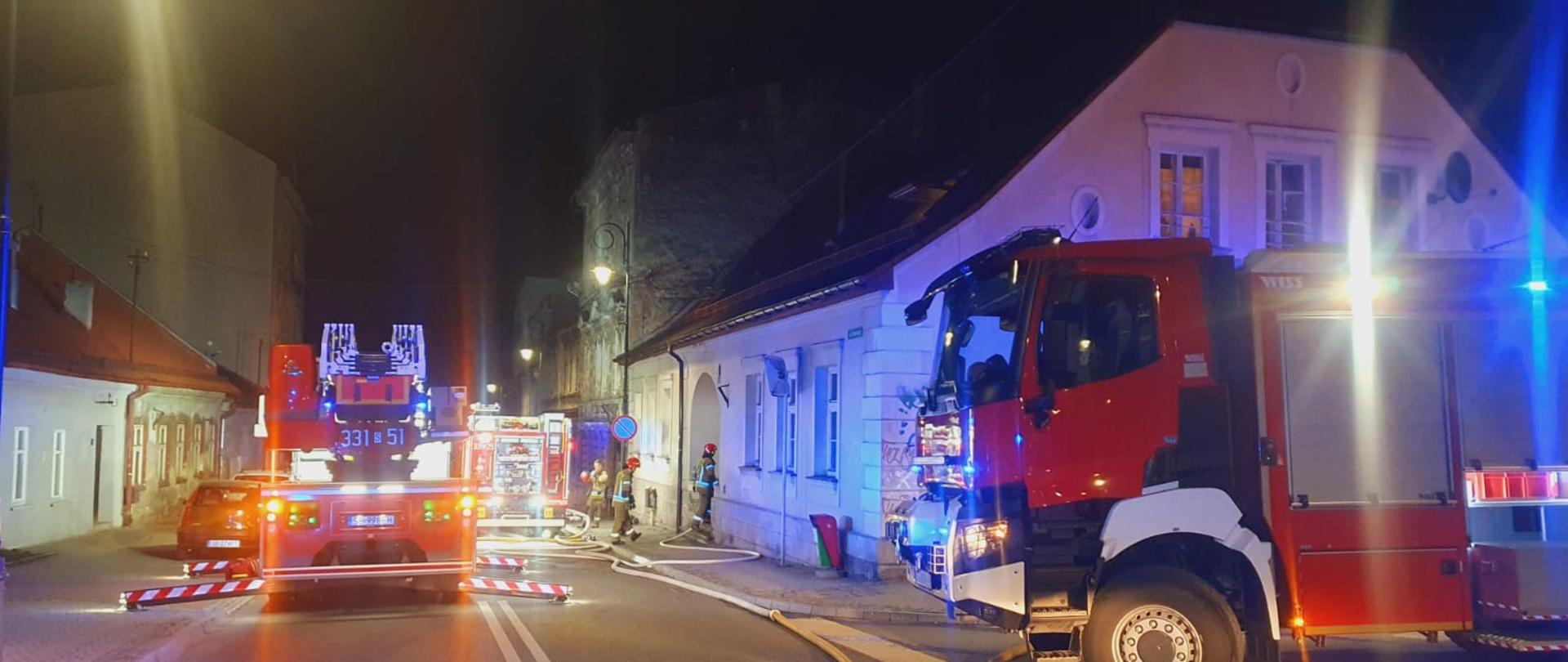 Zdjęcie wykonane na drodze jezdni podczas akcji ratowniczej. W lewym rogu zdjęcia znajduje się samochód pożarniczy z włączonymi sygnałami świetlnymi. Za pojazdem widoczny biały parterowy budynek mieszkalny. Z lewej strony zdjęcia widoczna drabina pożarnicza. W tle widoczni strażacy podczas działań ubrani w ubranie specjalne i hełm obok pojazdu pożarniczego.