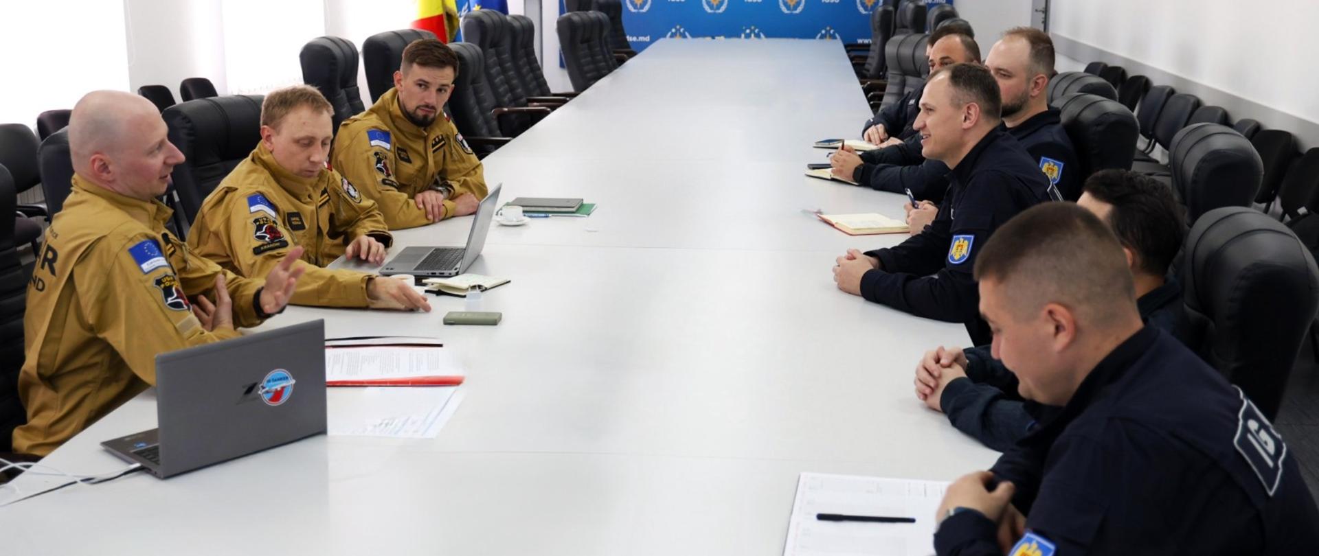 Strażacy siedzą przy długim stole konferencyjnym i prowadzą spotkanie robocze z przedstawicielami zagranicznej służby ratowniczej; na stole znajdują się laptopy i dokumenty.
