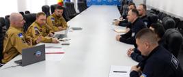 Strażacy siedzą przy długim stole konferencyjnym i prowadzą spotkanie robocze z przedstawicielami zagranicznej służby ratowniczej; na stole znajdują się laptopy i dokumenty.