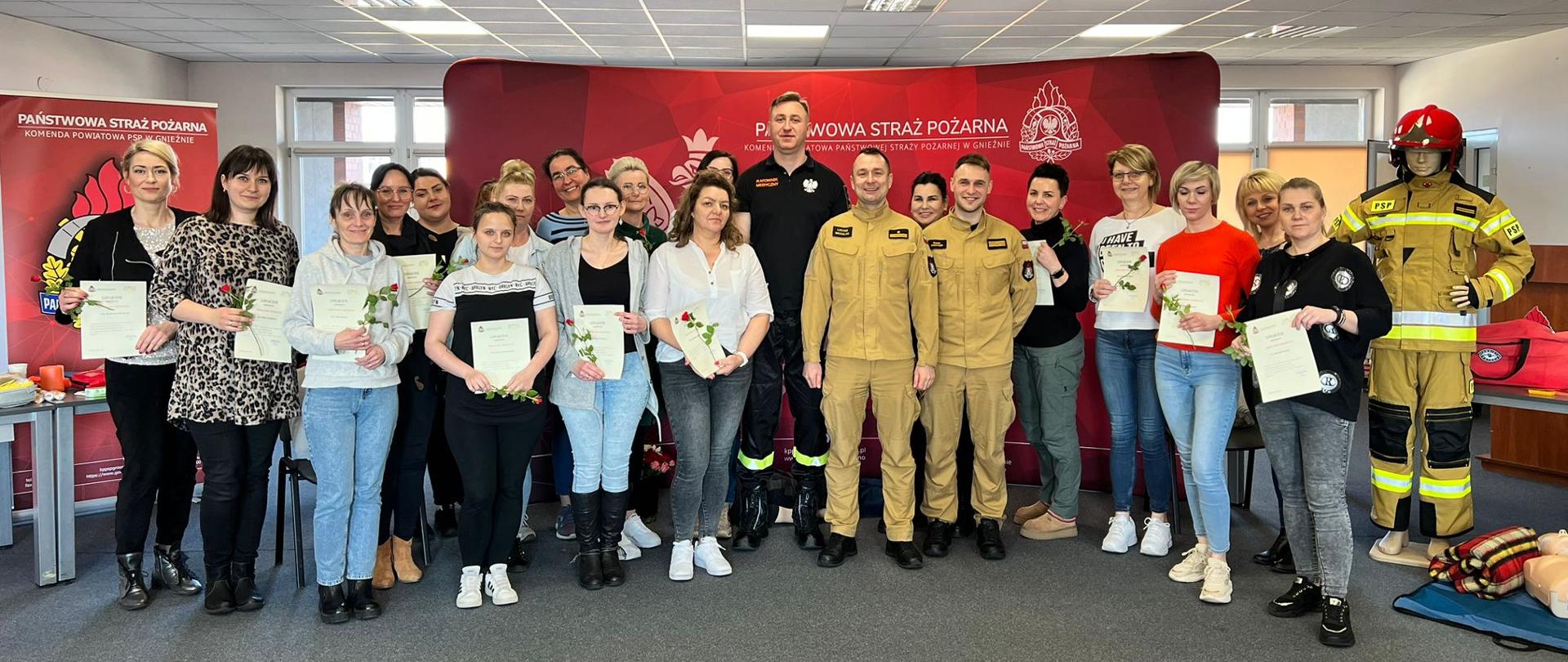 Grupa kobiet stoi ze strażakami trzymając kwiaty i dyplomy