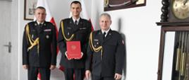 Trzech funkcjonariuszy Państwowej Straży Pożarnej stoi obok siebie strażak w środku trzyma czerwoną teczkę za nimi stoją dwie flagi Polski na ścianie wiszą obraz oraz szabla niedaleko stoi zegar.
