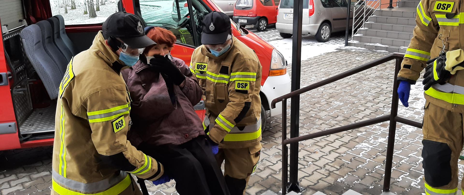 Fotografia przedstawiająca starszą kobietę przenoszoną przez dwóch strażaków ochotników do ZOZ na szczepienie w ramach Narodowego Programu Szczepień.