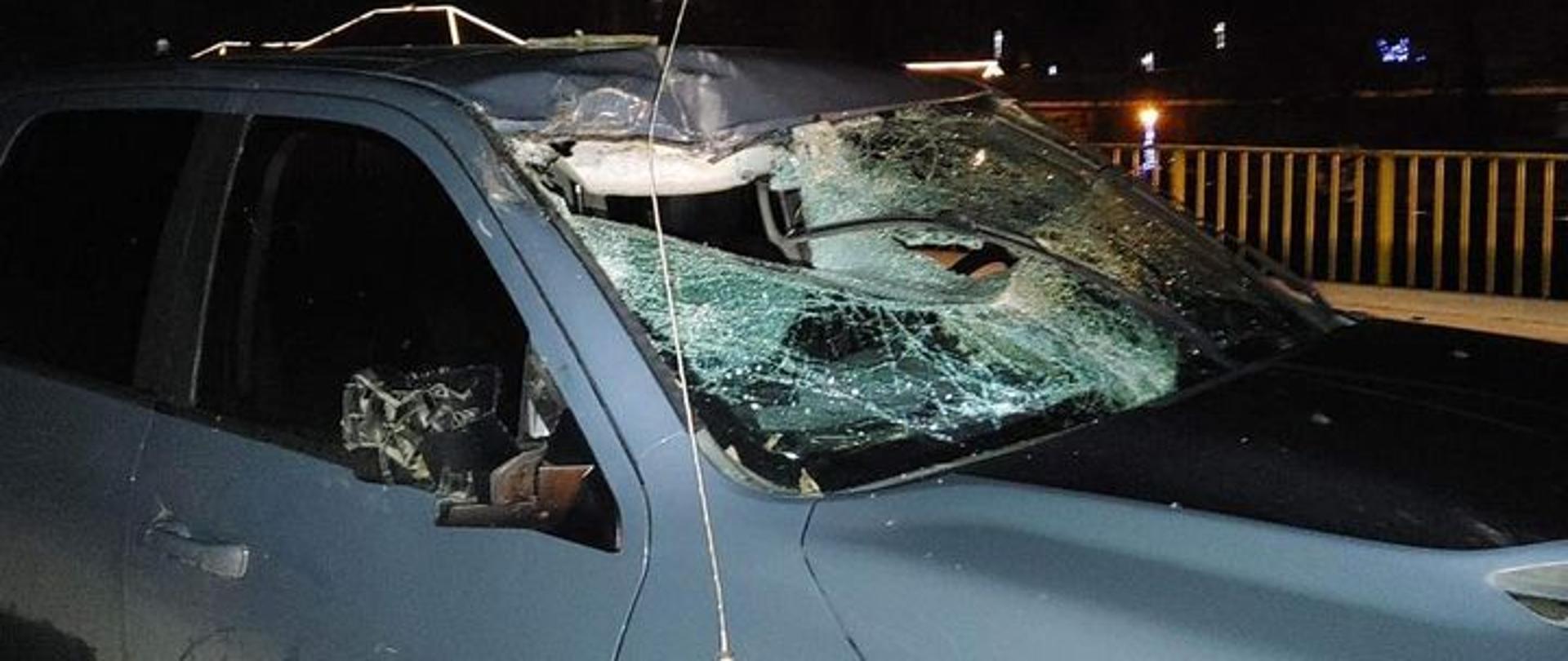 Samochód osobowy z uszkodzoną przednią szybą, wgniecionym dachem i połamanym prawym lusterkiem. 