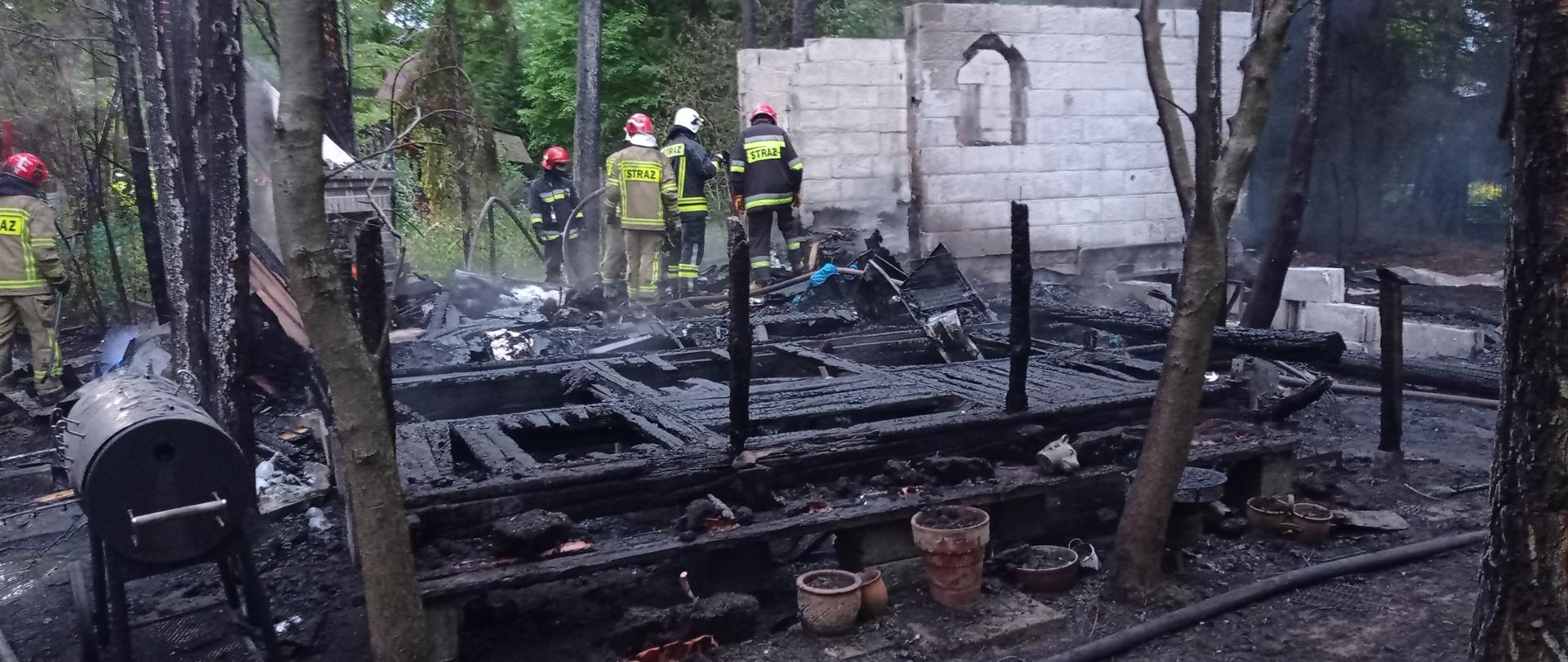 Zdjęcie przedstawia spalony budynek, tło drzewa iglaste. Budynek częściowo murowany z jasnych prefabrykatów. Na dole zdjęcia czarne spalone elementy drewniane konstrukcji budynku. Na pogorzelisku widać czterech strażaków dogaszających tlące się elementy. Strażacy ubrani w hełmy koloru czerwonego i białego, ubrania strażaków w kolorze piaskowym i granatowym. 