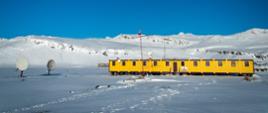 Widok na żółty parterowy budynek stacji, dookoła śnieg, za budynkiem niskie wzniesienia.