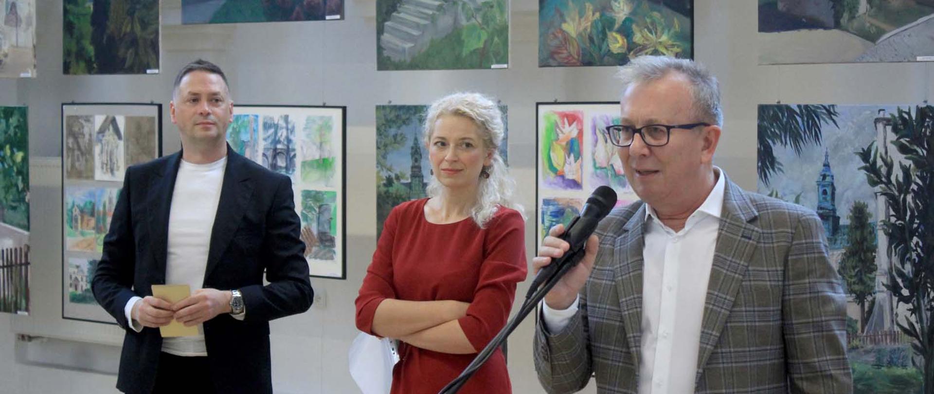 Zdjęcie przedstawia trzy osoby na tle ściany na której umieszczono barwne obrazy z pleneru.