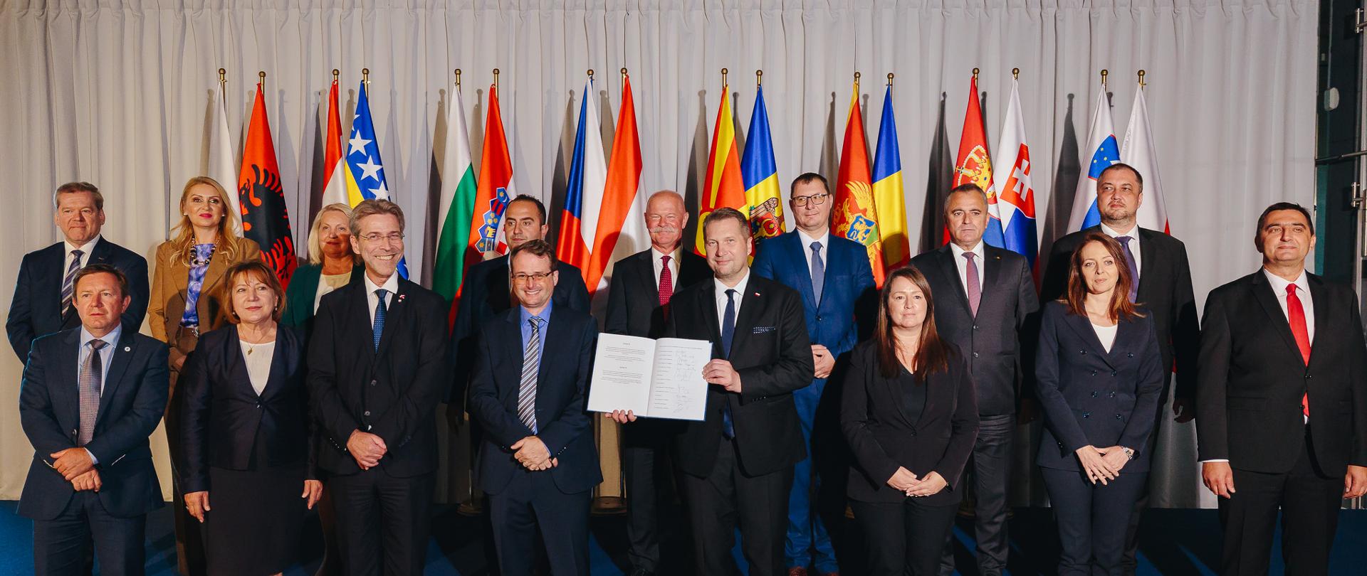 W dużej sali przed rzędem kolorowych flag stoi duża grupa elegancko ubranych osób, z przodu minister Czarnek trzyma w rękach otwarty dokument.

