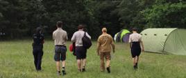 Po polanie idą w stronę namiotów obok siebie trzech harcerzy, strażak i policjantka.