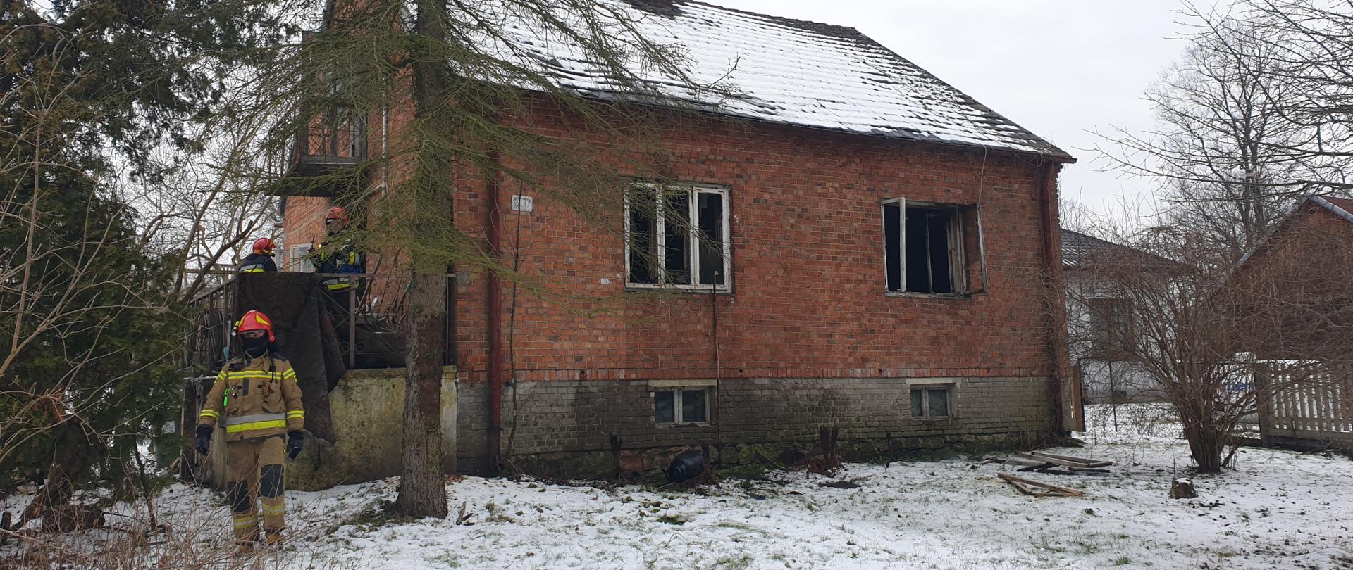 Dom jednorodzinny z cegły w którym doszło do pożaru. Okna w budynku otwarte. Po lewej stronie strażak w umundurowaniu bojowym koloru piaskowego.