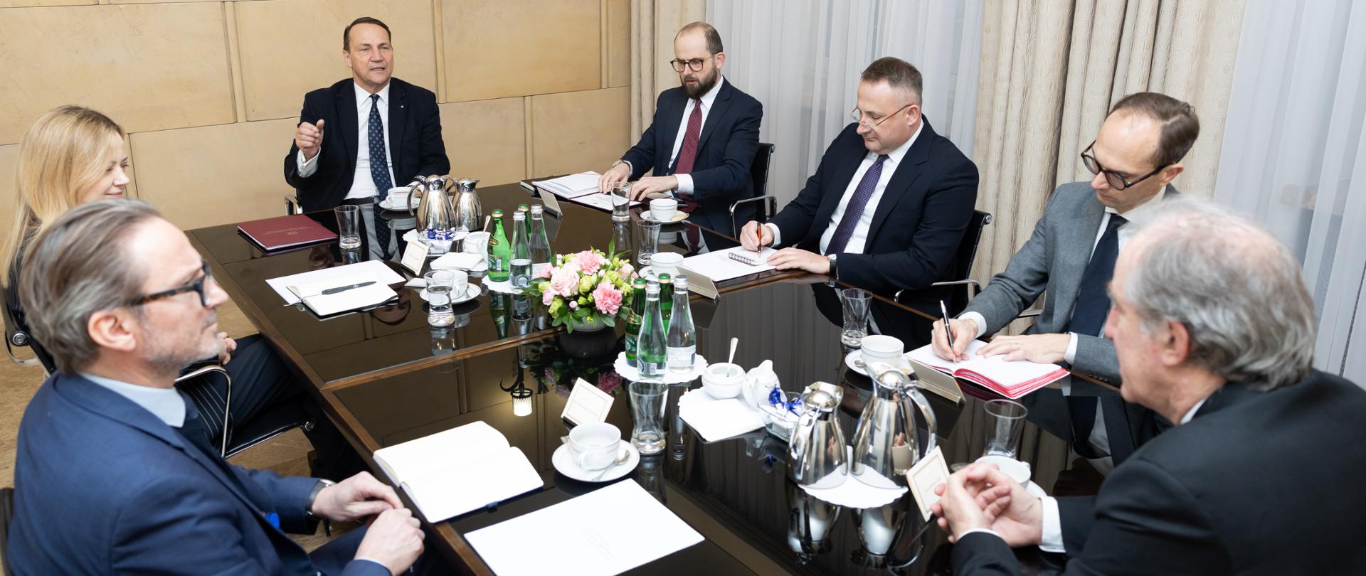 6 mężczyzn i jedna kobieta siedzący przy stole i rozmawiający podczas spotkania.