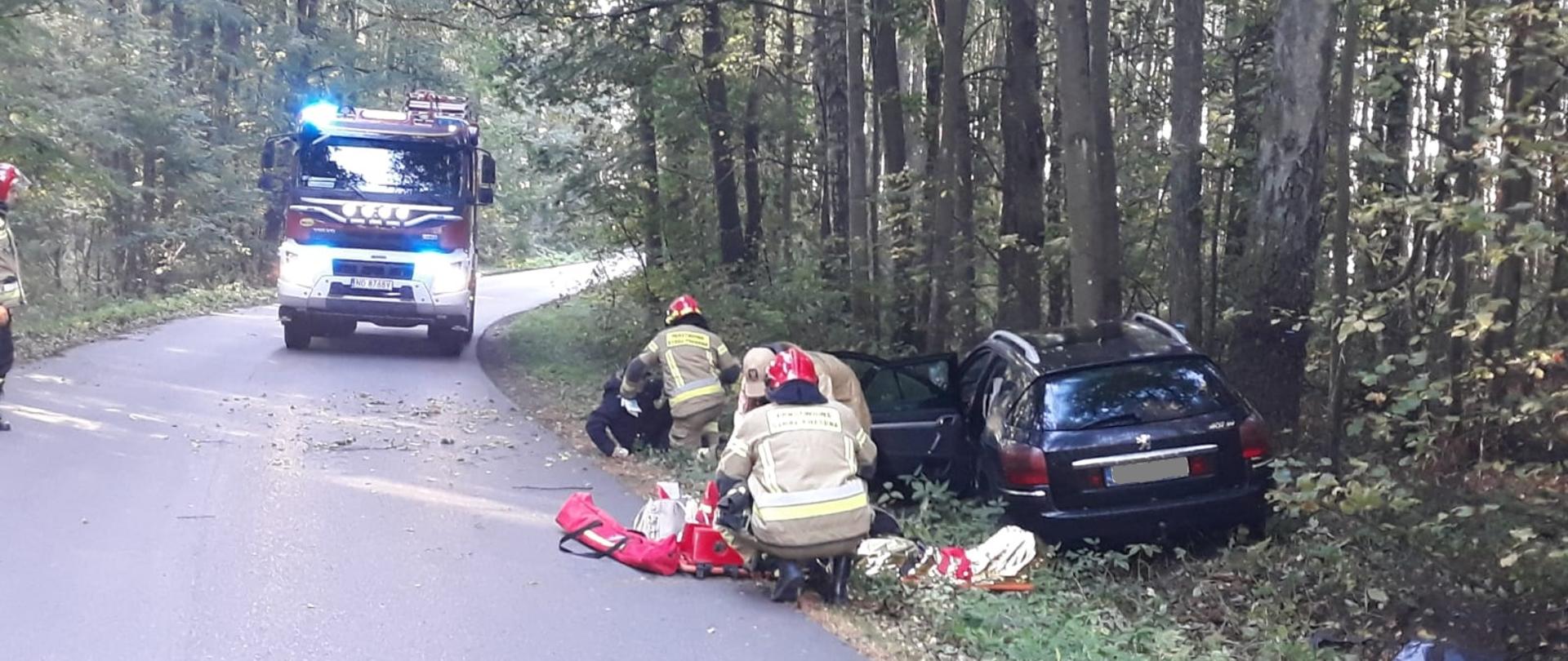 Zdjęcie przedstawia strażaków udzielających pomocy osobie poszkodowanej, w tle rozbity samochód oraz pojazd pożarniczy