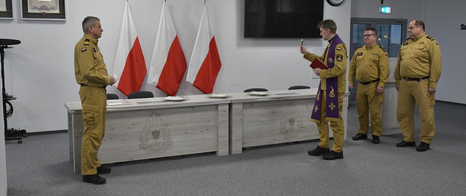 Kapelan pomorskich strażaków święci opłatek położony na biurkach po dwóch stronach stoi kierownictwo Komendy Wojewódzkiej Państwowej Straży Pożarnej w Gdańsku za biurkami ustawione są trzy flagi Polski na ścianie wisi orzeł telewizor zegar oraz rota ślubowania. 