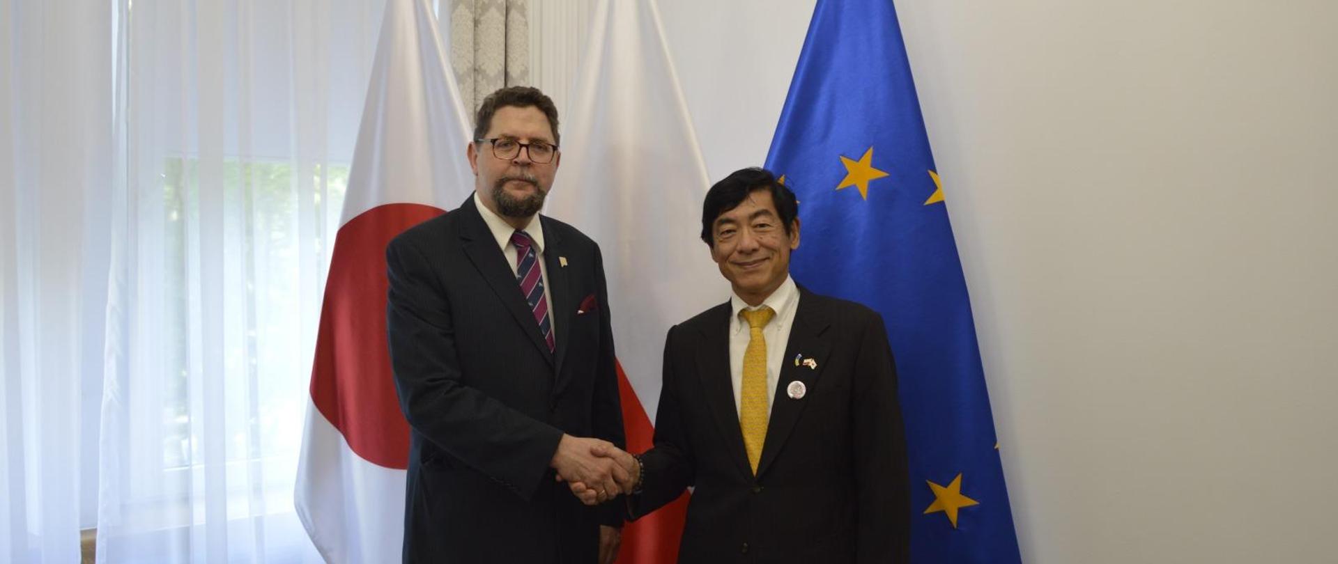 Pod ścianą przy której stoją flagi Polski i UE stoi wiceminister Szeptycki który podaje rękę mężczyźnie w czarnym garniturze.