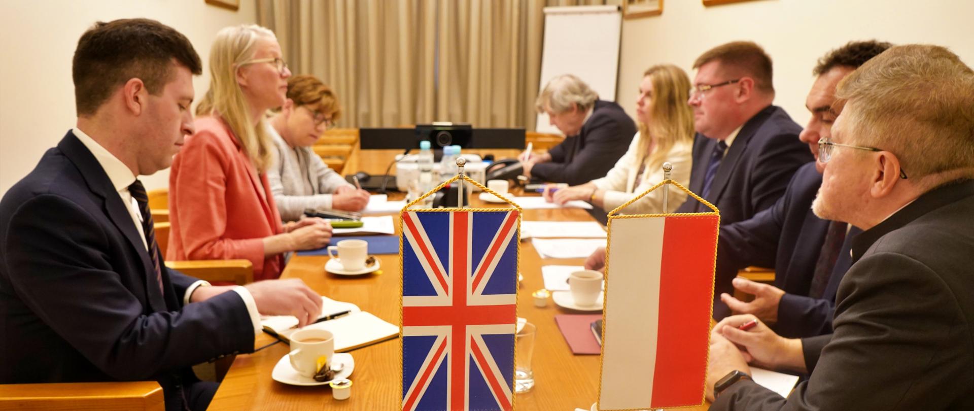 Na środku stół. Po prawej dwie kobiety i mężczyzna po lewej czterech mężczyzn i kobieta. Na stole filiżanki oraz kartki papieru i dwie flagi: polska i brytyjska