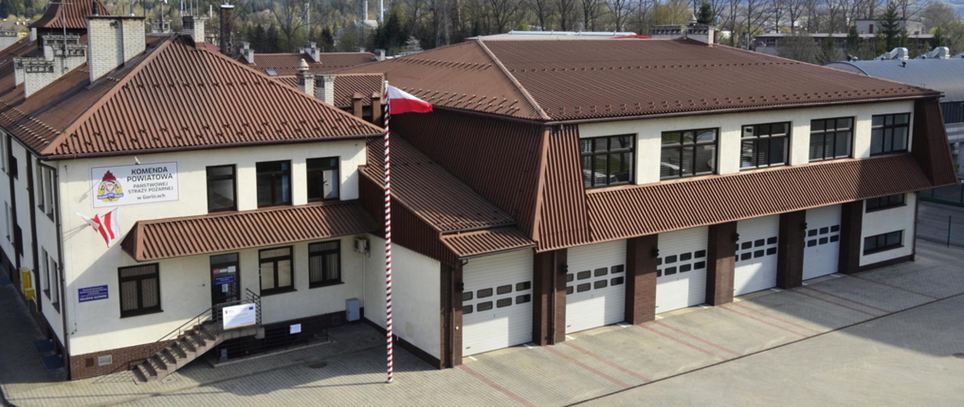 Na zdjęciu z lotu ptaka widoczny budynek KP PSP w Gorlicach. Brązowy dach biała elewacja. Przed budynkiem plac oraz maszt z powiewającą flaga biało czerwoną.