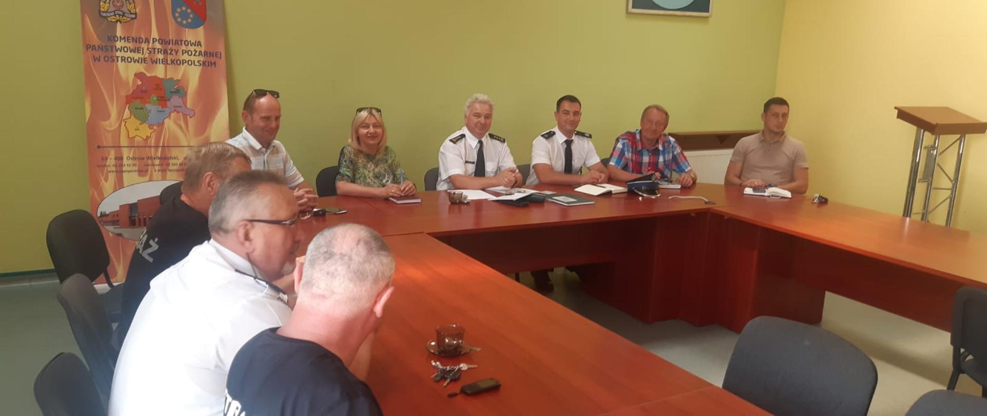 Funkcjonariusze i ochotnicy siedzący na sali narad w podkowie, W samym środku komendant.