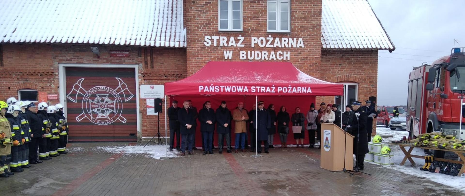 Na placu przed OSP stoi namiot pod, ktoym są goście po leweij stronie stoją pododziały a po prawej samochód gasniczy obok, którego leży sprzęt do przekazania. Pora zminowa. 