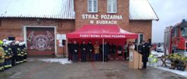 Na placu przed OSP stoi namiot pod, ktoym są goście po leweij stronie stoją pododziały a po prawej samochód gasniczy obok, którego leży sprzęt do przekazania. Pora zminowa. 