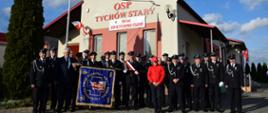 Na tle żółto-czerwonej remizy z dużym czerwonym napisem na elewacji OSP Tychów Stary druhowie OSP prezentują nowy sztandar wraz z zaproszonymi gośćmi. Wszyscy w strojach galowych