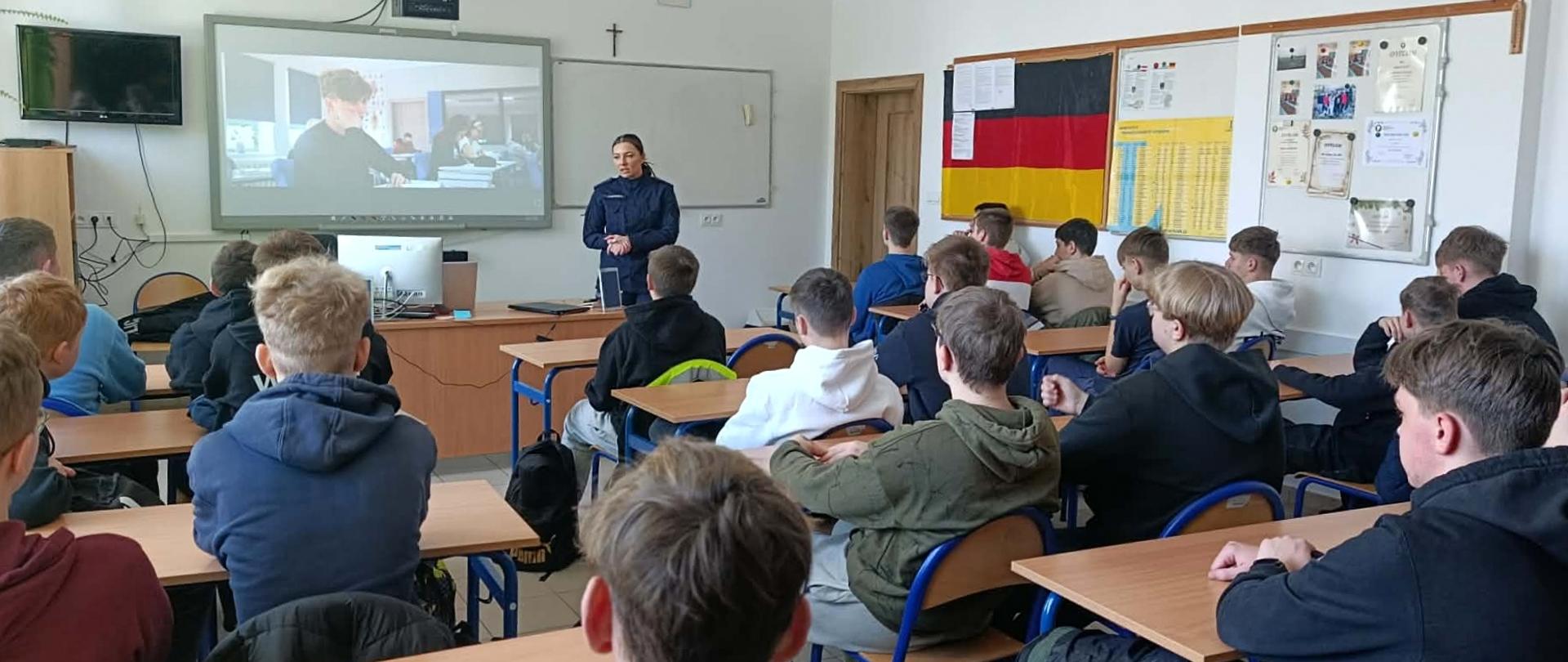 Policjantka prowadzi prelekcję dla uczniów siedzących w ławkach w sali lekcyjnej wyposażonej w tablicę interaktywną z wyświetlonym filmem.