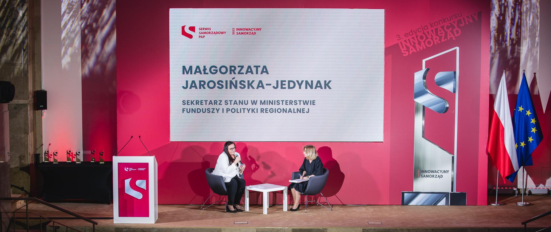 W sali konferencyjnej na scenie dwie kobiety rozmawiają przy stoliku. Z lewej wiceminister funduszy i polityki regionalnej Małgorzata Jarosińska-Jedynak.
