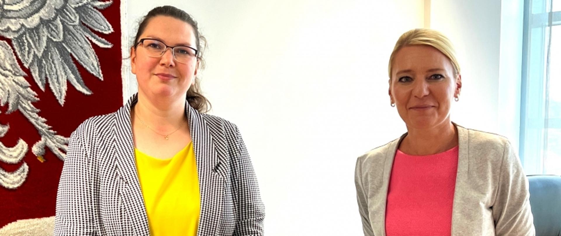 Dwie osoby w marynarkach na tle czerwonej dekoracji z białym wzorem piór lub liści.