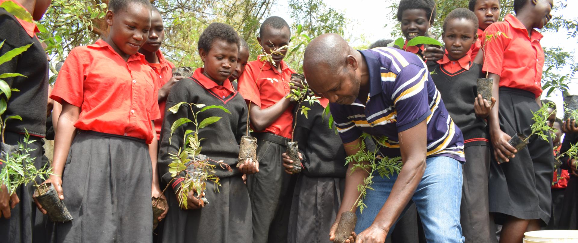 Grupa uczniów trzymających w rękach sadzonki przygląda się nauczycielowi sadzącemu drzewko