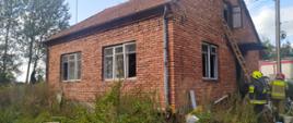 Na zdjęciu jest dom parterowy z czerwonej cegły, przy wejściu do domu strażacy, w tle widać kawałek wozu strażackiego, przed domem panuje nieład. 
