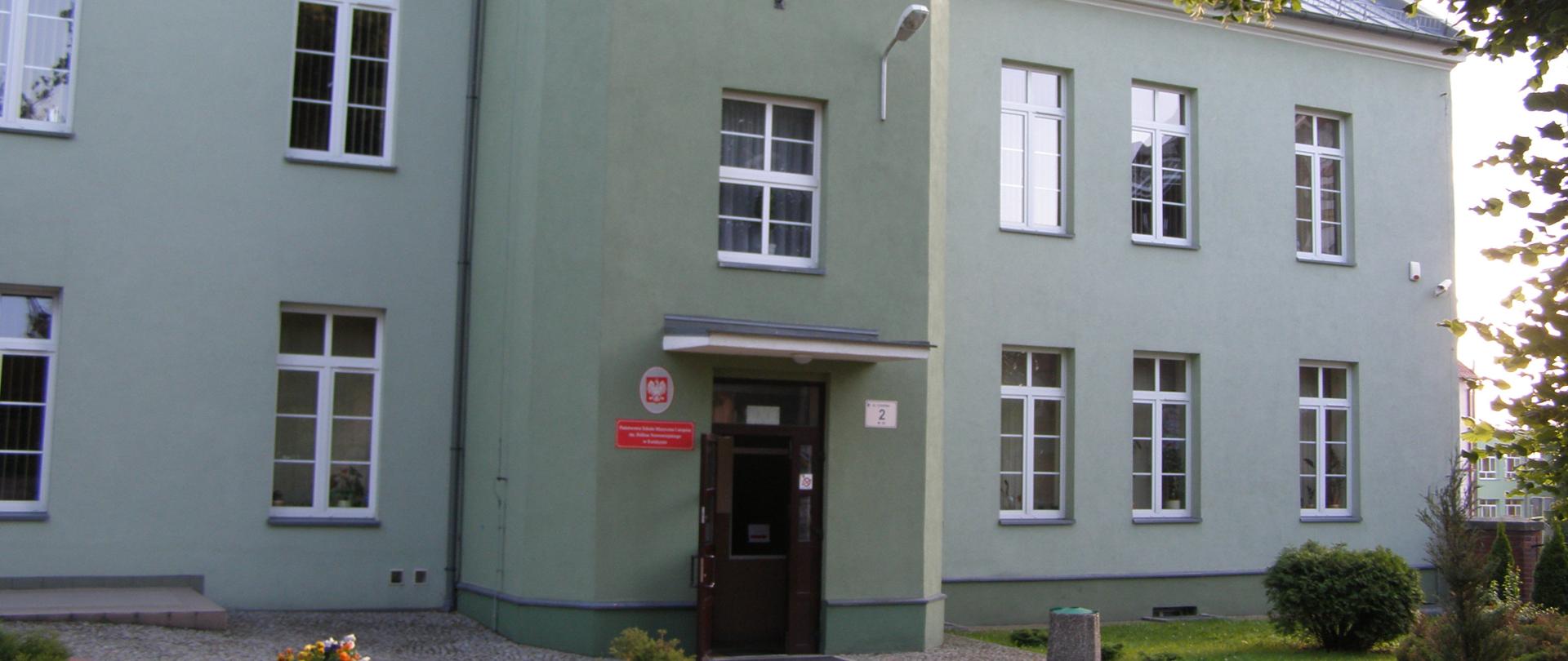  Zielony, dwupiętrowy budynek szkoły z czerwoną tablicą nazwy szkoły po lewej stronie wejścia głównego