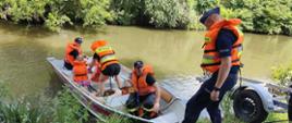 Strażacy wodują łodź ratowniczą w której są ratownicy oraz dwójka dzieci. Na brzegu stoi policjant. 