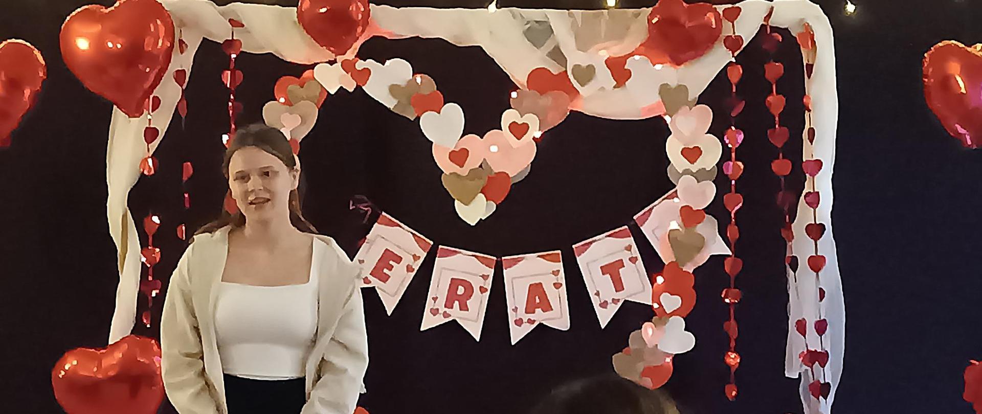 Na środku, przed czarnym tłem z lampkami, stoi młoda kobieta w jasnej bluzce, jasnej rozpinanej bluzie i czarnych spodniach. Za nią znajduje się dekoracyjna brama ozdobiona białą tkaniną oraz papierowymi sercami. Wokół wiszą liczne czerwone balony w kształcie serc oraz girlandy z serduszek. Na pierwszym planie widać siedzących uczestników wydarzenia, a po prawej stronie stół nakryty biało-czerwoną, kraciastą serwetą z czerwonymi dekoracjami.