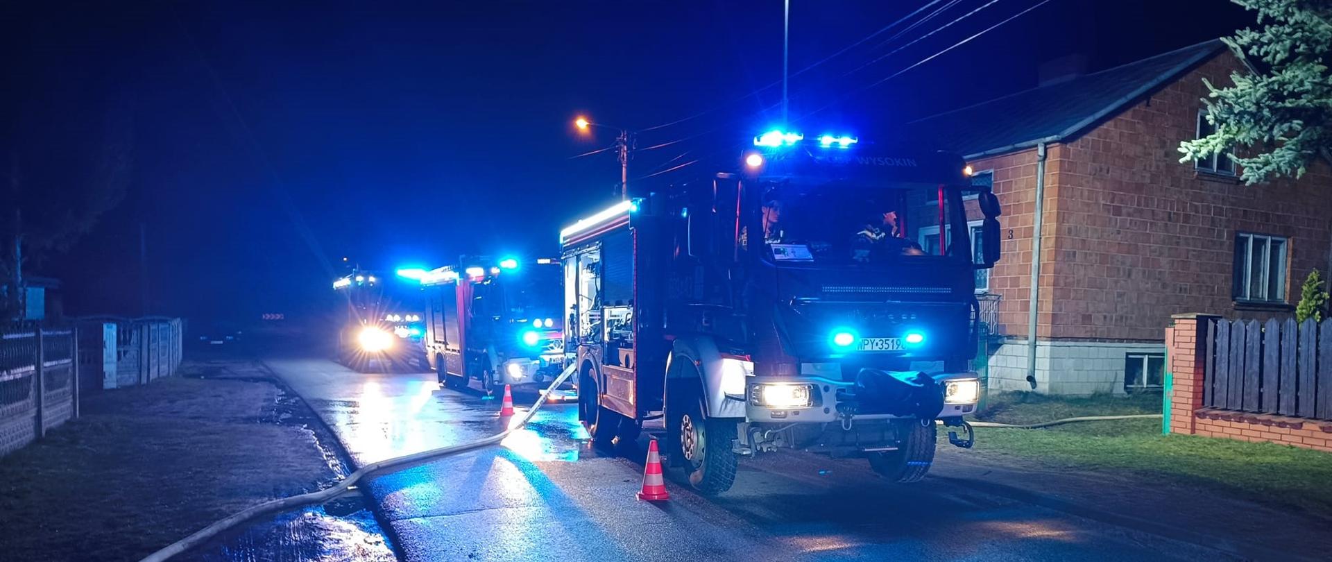 Zdjęcie przedstawia scenę nocną z trzema wozami strażackimi stojącymi na ciemnej, mokrej drodze, co sugeruje akcję gaśniczą. Wozy strażackie mają włączone niebieskie światła sygnalizacyjne, które intensywnie oświetlają otoczenie i nadają scenie dramatyczny charakter. Na pierwszym planie, po prawej stronie, widać przód dużego wozu strażackiego. Przed nim i koło niego na asfalcie stoją trzy pachołki drogowe w czerwono-białe pasy. Od wozu biegnie wąż strażacki, który leży na drodze i jest częściowo widoczny po lewej stronie kadru. Za pierwszym wozem stoją dwa kolejne, również z włączonymi światłami.
W tle, po prawej stronie, widoczny jest fragment budynku mieszkalnego z ceglaną fasadą oraz fragment ogrodzenia. Po lewej stronie widać fragmenty płotu i inne budynki w oddali, ginące w ciemności. Niebo jest całkowicie czarne, co potęguje wrażenie nocy.