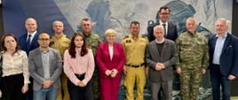 Na zdjęciu przedstawiono uczestników wizyty pozujących do wspólnej fotografii na tle ścianki z logo Polskiej Grupy Zbrojeniowej (PGZ). W centrum kadru stoją posłowie, przedstawiciele administracji publicznej, kierownictwa instytucji oraz służb mundurowych. Wśród zgromadzonych znajdują się funkcjonariusze Państwowej Straży Pożarnej w piaskowych mundurach służbowych oraz żołnierze w mundurach wojskowych w kamuflażu. Pozostali uczestnicy ubrani są w stroje biznesowe i formalne. W tle widoczna jest duża plansza z logo PGZ, wskazująca na związek spotkania z tematyką bezpieczeństwa i obronności. Zdjęcie wykonano wewnątrz budynku, w dobrze oświetlonym pomieszczeniu podczas oficjalnej części wizyty.