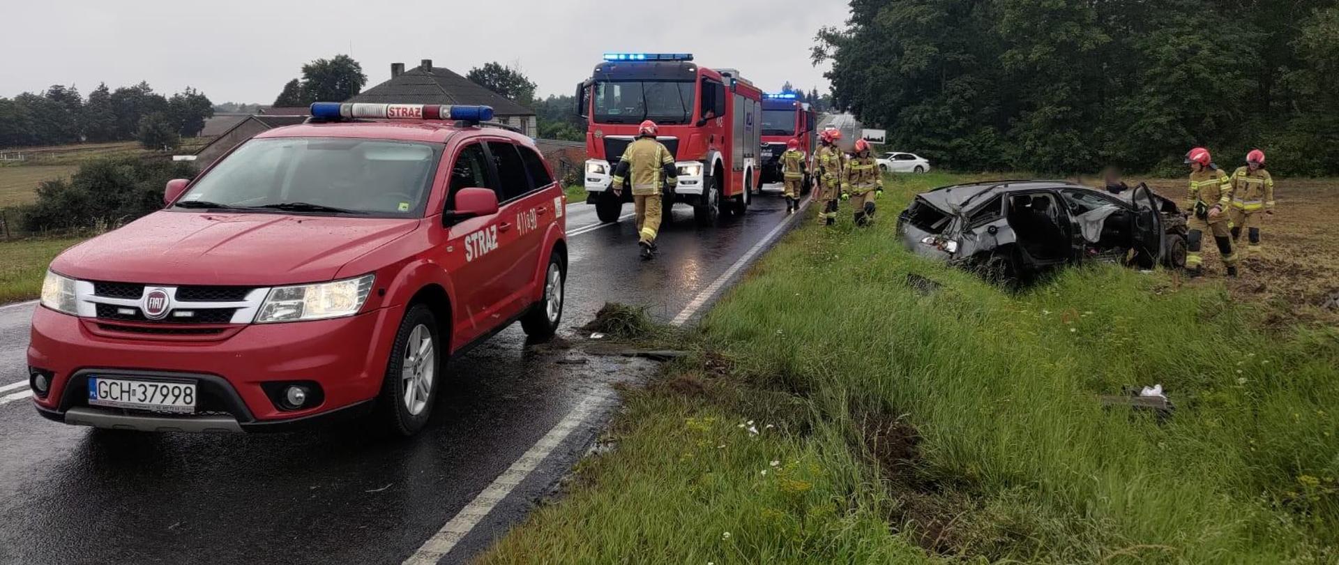 Deszczowy dzień na drodze zaparkowane trzy czerwone samochody straży pożarnej, jako pierwszy osobowy a za nim dwa ciężarowe. Samochody mają włączone światła mijania oraz niebieskie ostrzegawcze. W rowie leży wrak czarnego samochodu osobowego. Na terenie akcji widać pracujących strażaków ubranych w piaskowe ubrania specjalne i czerwone hełmy. 