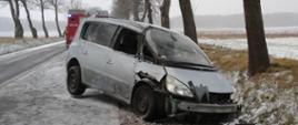 Uszkodzony samochód osobowy stojący częściowo w przydrożnym rowie, częściowo na pasie jezdni. Jezdnia i pobocze pokryte cienką warstwą śniegu. W tle wóz strażacki.