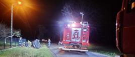 Noc, widok na wypadek komunikacyjny, na pierwszym planie widoczny wóz straży pożarnej oświetlający miejsce wypadku, po prawej stronie widoczny znajdujący się poza drogą przewrócony na bok samochód osobowy, za autem grupa strażaków prowadzi działania ratownicze, udzielające pierwszej pomocy poszkodowanej osobie