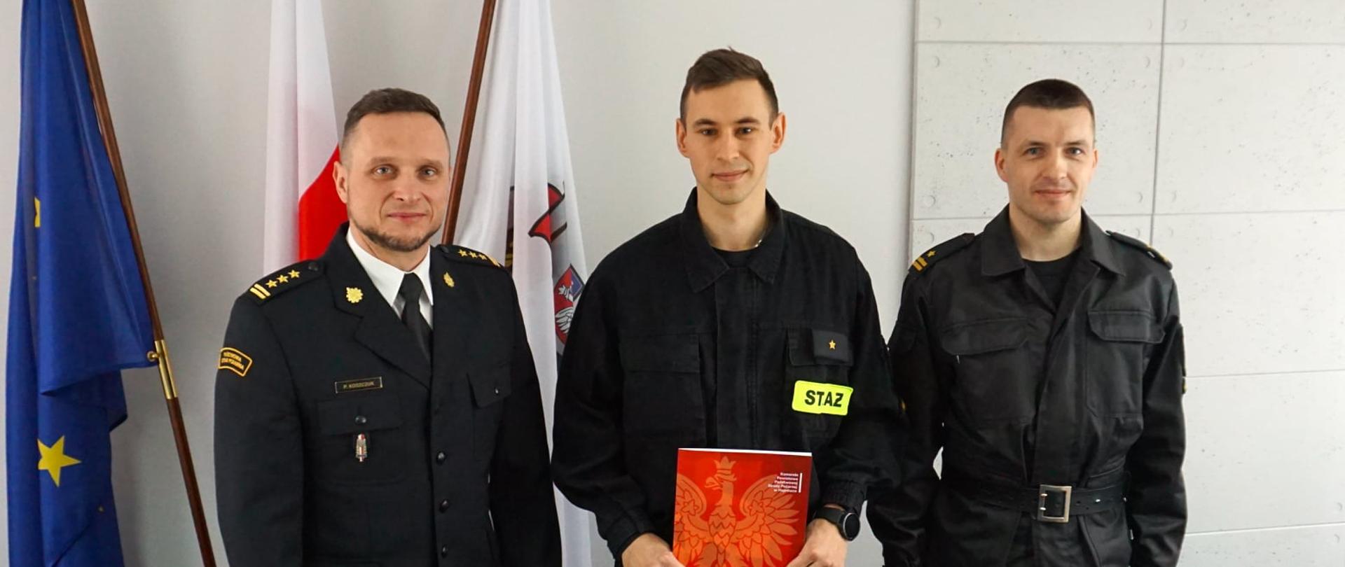 W pomieszczeniu na tle fag: Polski UE oraz flagi z logo PSP stoi 3 strażaków w czarnych mundurach.