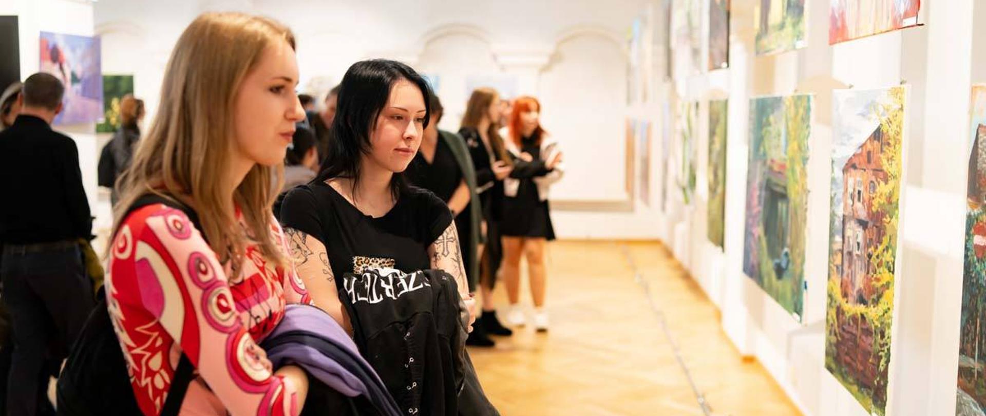 Na pierwszym planie widać dwie kobiety oglądające obrazy, które zajmują prawą część zdjęcia. W tle widać inne osoby oglądające malarstwo. 