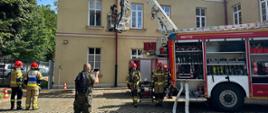 Zdjęcie przedstawia strażaka próbującego dotrzeć przy pomocy drabiny mechanicznej do osoby poszkodowanej, stojącej w konie na pierwszym piętrze budynku.