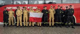 Na zdjęciu znajduje się grupa strażaków w ubraniach koszarowych. Pozują do zdjęcia grupowego na tle autobusu strażackiego. W środku grupy dwie osoby trzymają flagę Polski.