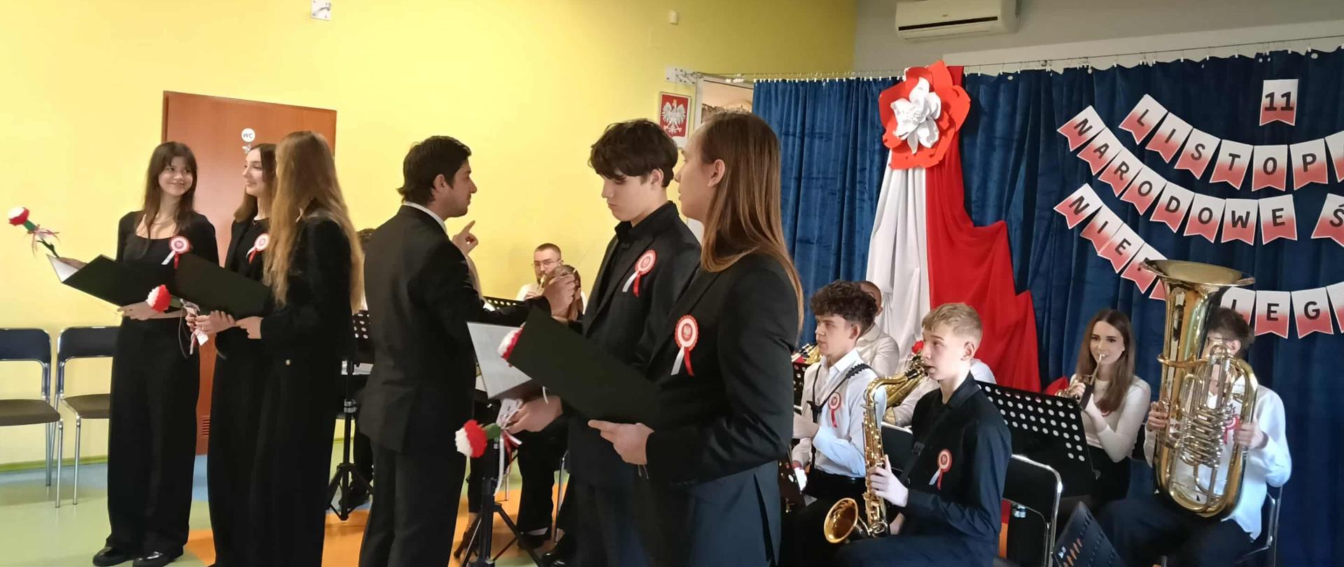 Uczniowie i nauczyciele występują podczas koncertu patriotycznego w Ośrodku Rehabilitacyjno-Edukacyjno-Wychowawczym w Mielcu; młodzież śpiewa i gra na instrumentach, a sala udekorowana jest biało-czerwonymi barwami