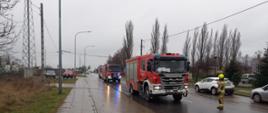 Zdjęcie wykonane w ciągu dnia. Zachmurzenie duże. Widać samochody ratowniczo-gaśnicze. Pojazdy są w kolorze czerwonym. Posiadają sygnalizację ostrzegawczą. Zdjęcie wykonane na drodze publicznej.