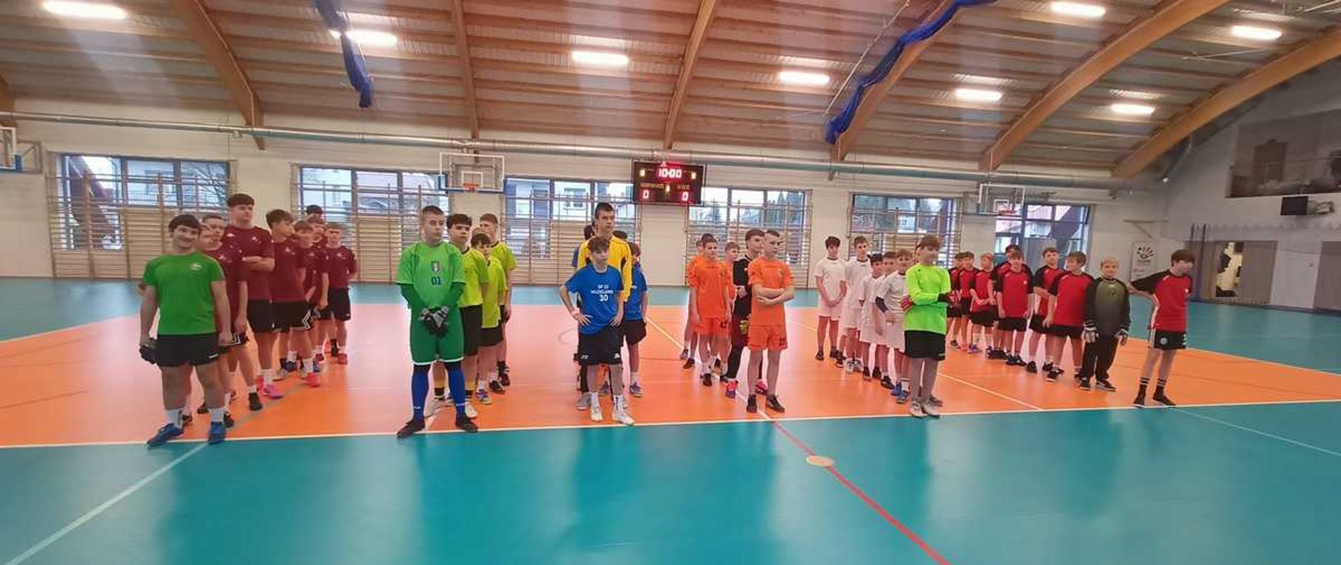 Na zdjęciu widać sześć drużyn chłopców ustawionych w równych szeregach, jak przed rozpoczęciem zawodów. Każda grupa ma inne stroje.