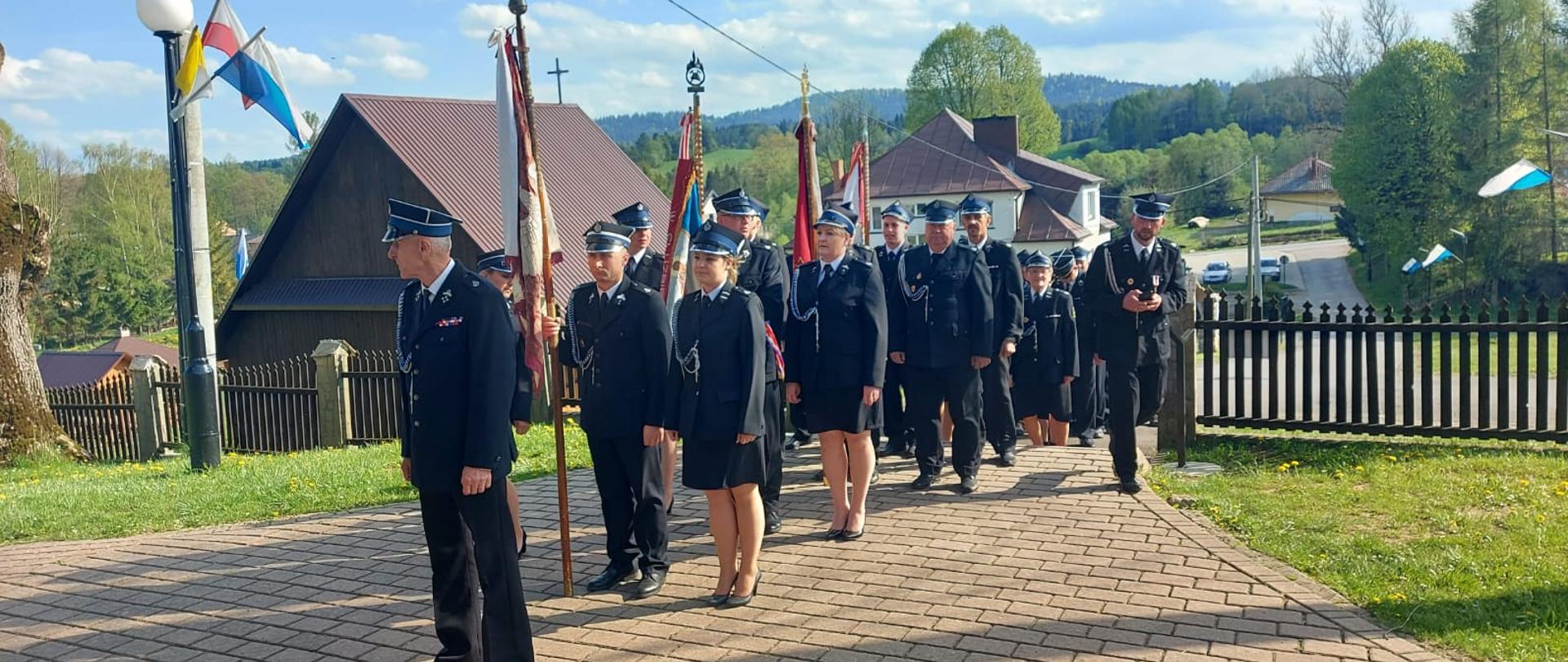 Na zdjęciu znajdują się strażacy OSP w mundurach galowych trzymający sztandary. Za nimi domy.