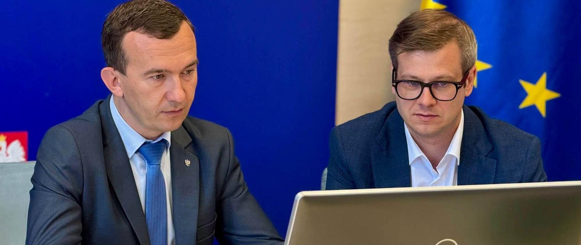 2 mężczyzn w garniturach, wojewoda oraz dyrektor wydziału polityki społecznej, siedzą w sali konferencyjnej, patrzą na 1 monitor laptopa.
