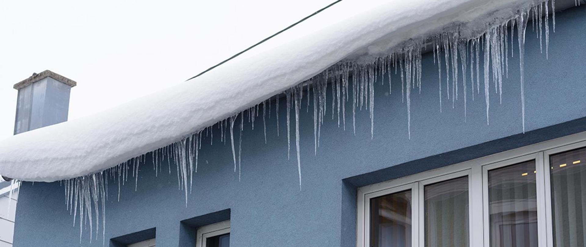 Zdjęcie przedstawia śnieg zalegający na dachu i zamarznięte sople lodu zwisające z dachu