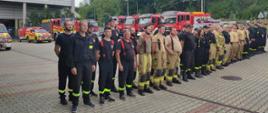 Na zdjęciu widoczni strażacy Ochotniczych Straży Pożarnych ubrani w ubrania piaskowe i czarne uczestniczący w podsumowaniu powiatowych ćwiczenia taktyczno-bojowe pod kryptonimem. „Empol 2023” na placu zewnętrznym Komendy powiatowej Państwowej Straży Pożarnej w tle czerwone samochody Pożarnicze.