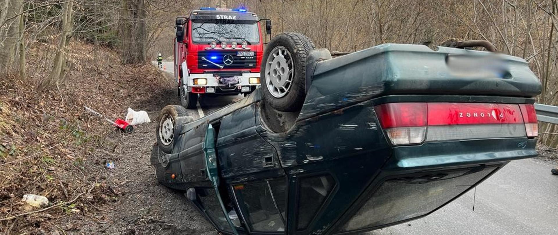 Samochód osobowy na dachu a w tle samochód pożarniczy.