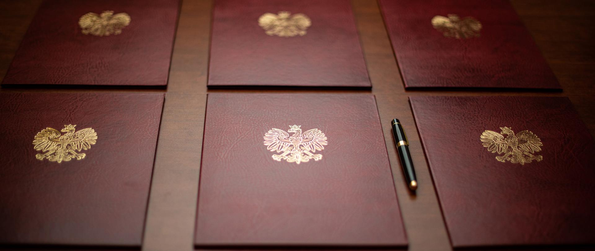 Na zdjęciu widać sześć ułożonych twardych okładek na dokumenty z wytłoczonym orłem z godła narodowego. Obok jednej z teczek leży eleganckie pióro do pisania.