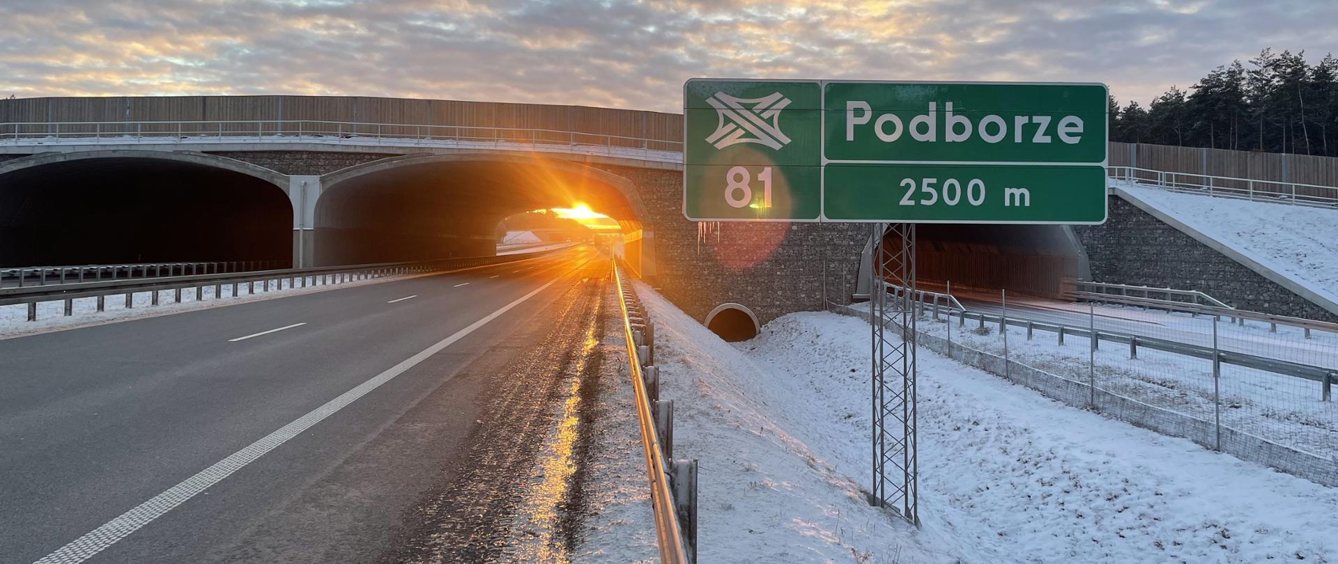 Droga ekspresowa biegnąca aż pod horyzont, na pierwszym planie tablica informująca o najbliższym węźle drogowym Podborze, za nią przejście dla zwierząt nad drogą, zachodzące słońce